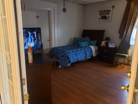 A small bedroom with a single bed covered in blue patterned bedding and multiple pillows. There is a dark wooden nightstand with a lamp, books, and a teddy bear next to the bed. A chair is placed near a window with brown curtains. A TV is on a dresser opposite the bed, and a door labeled '02' is visible in the background. The room has wooden flooring and light-colored walls.