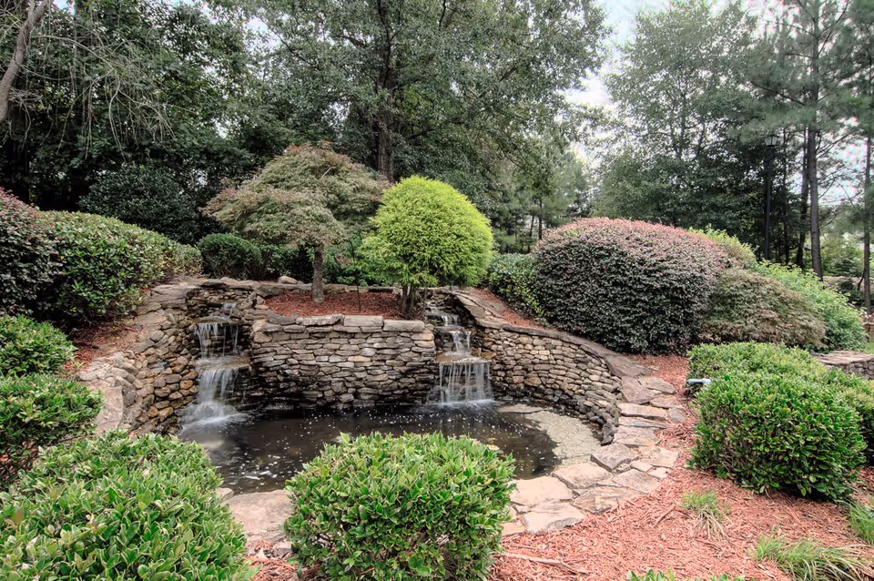 A serene outdoor garden area featuring a small stone waterfall flowing into a pond, surrounded by various green shrubs, bushes, and trees with a mulched ground.