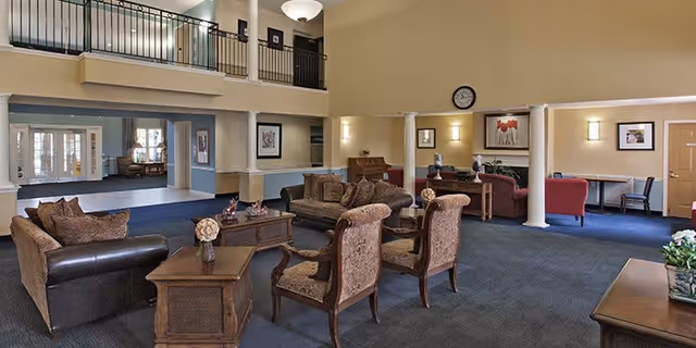 A spacious living room area in a senior living facility with high ceilings and a balcony railing above. The room is furnished with multiple upholstered chairs and sofas arranged around wooden coffee tables. The walls are painted beige with framed artwork and a clock. There are columns dividing the space and a piano against the far wall. The carpet is blue, and there is natural light coming from windows in the background.