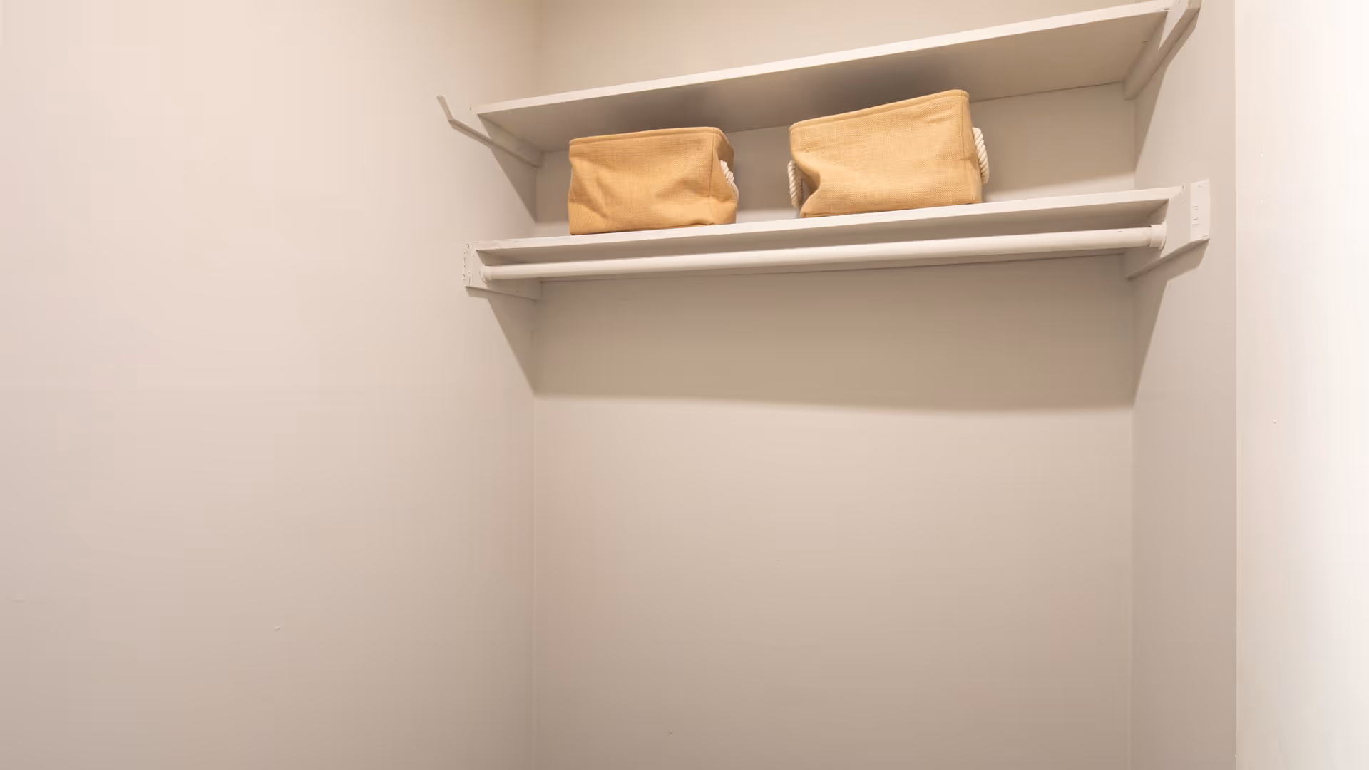 Empty closet with a white shelf and hanging rod, containing two beige fabric storage bins on the shelf.