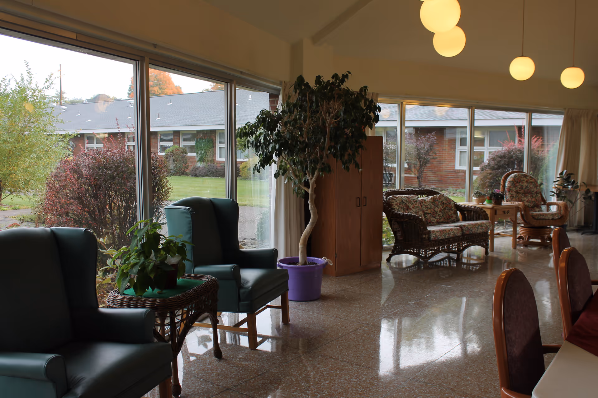 A bright and spacious common area with large floor-to-ceiling windows showing a garden outside. Inside, there are comfortable armchairs, wicker chairs with floral cushions, a small table with a potted plant, a tall potted tree in a purple pot, and a wooden cabinet. The floor is shiny and reflective, and round ceiling lights hang above.