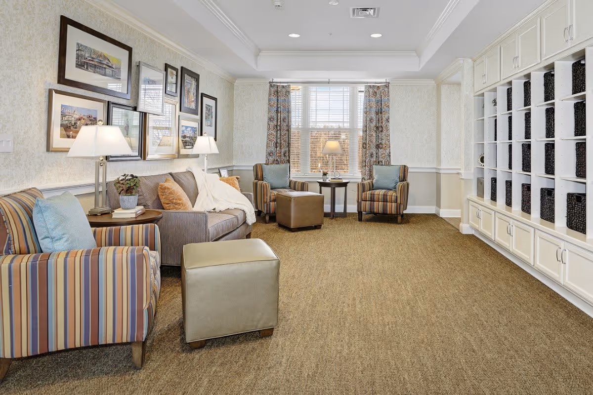 Sunlit communal lounge with sofas and striped armchairs arranged around ottomans, lamps, wall art, and built-in cubby shelving.