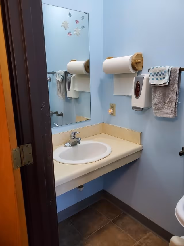 Small bathroom with light blue walls, a beige countertop with a white sink and silver faucet, a large mirror with decorative stickers, a paper towel roll mounted on the wall, a soap dispenser, and a towel hanging on a rack.