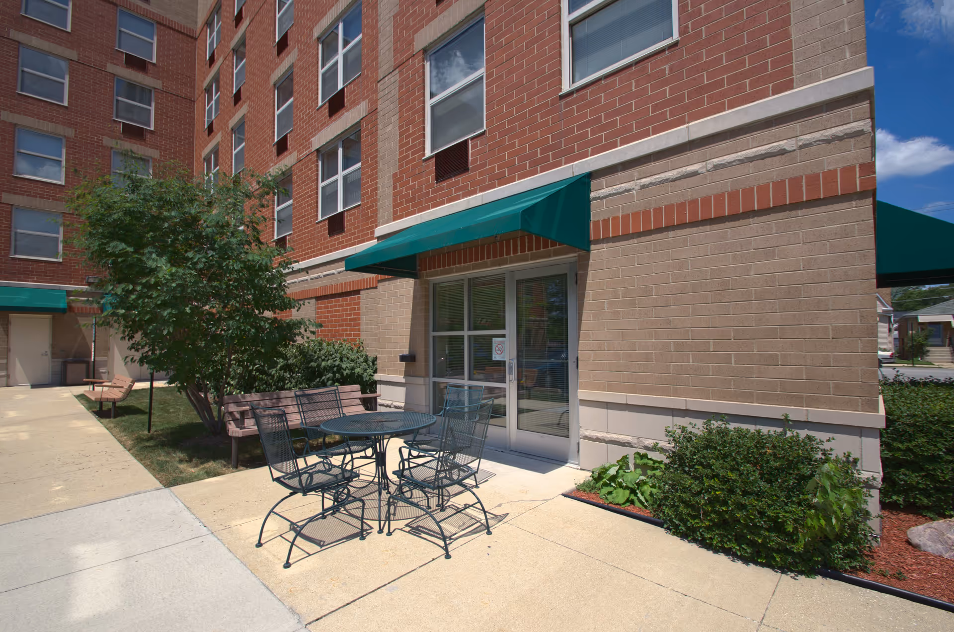 Outdoor patio area at Senior Suites of Jefferson Park featuring a round metal table with four matching chairs, two wooden benches, green bushes, and a tree next to a brick building with multiple windows and green awnings above the doors.