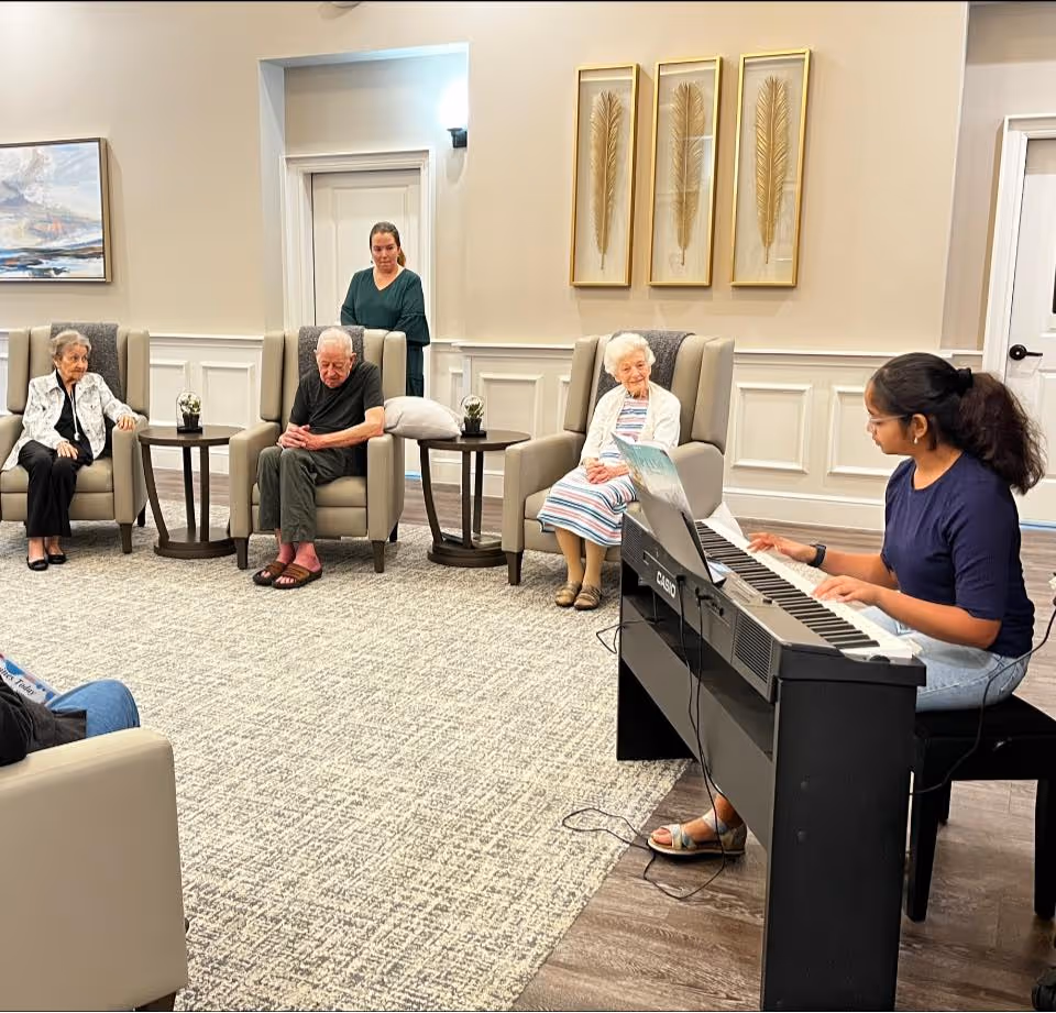 A young woman plays a keyboard piano in a cozy living room setting while three elderly people sit in armchairs listening. A staff member stands in the background near a doorway. The room has light-colored walls, decorative framed feathers, and a carpeted floor.