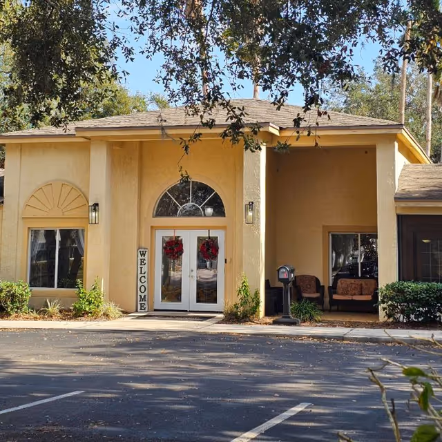 Front exterior view of a beige senior living facility building with a double glass door entrance decorated with red wreaths. There is a vertical 'WELCOME' sign next to the door, two wall-mounted lights, and a small seating area with two chairs and a table on the right side. Trees and bushes surround the building, and a parking lot is visible in the foreground.