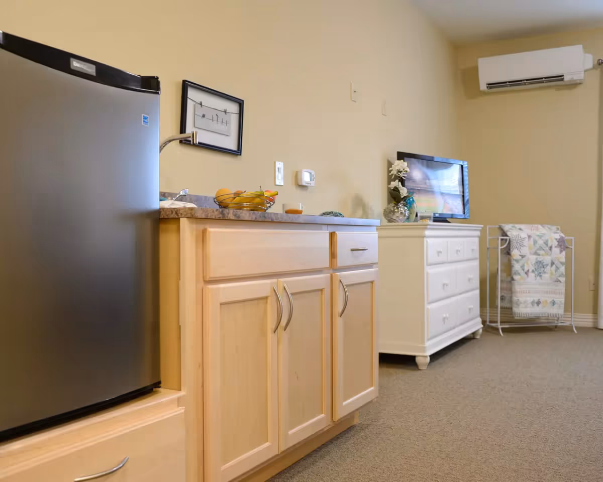 Interior view of a room featuring a small stainless steel refrigerator next to light wood cabinets with a countertop holding a fruit basket. On the wall above the counter is a framed picture. In the background, there is a white dresser with a flat-screen TV on top, a decorative vase with flowers, and a quilt rack holding a patterned quilt. An air conditioning unit is mounted on the wall above the dresser.