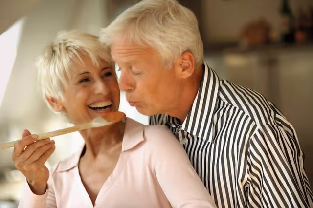 An elderly couple in a kitchen setting, the woman holding a wooden spoon with food on it while the man leans in to taste it, both smiling and enjoying the moment.