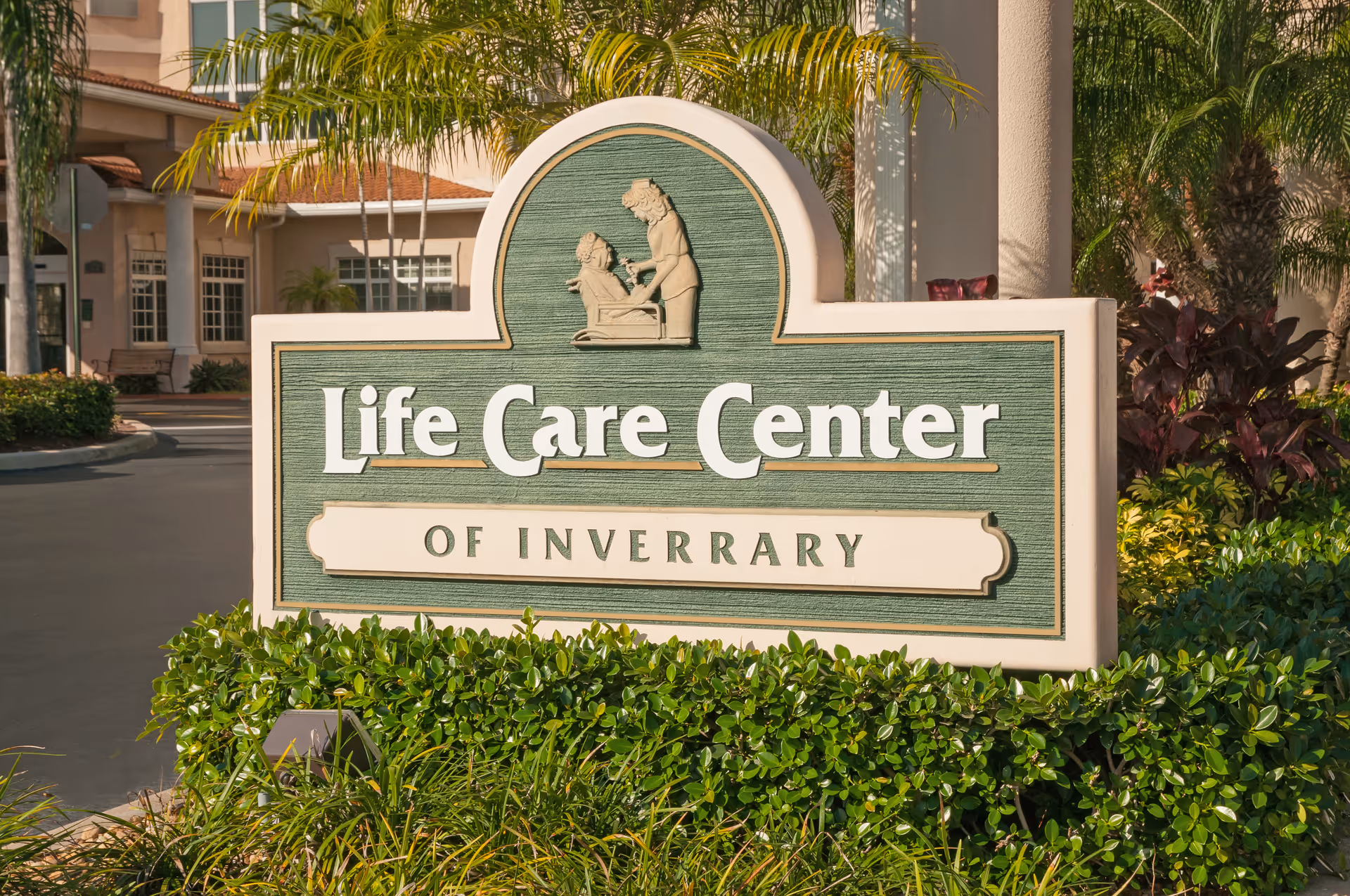 A large green and beige sign reading 'Life Care Center of Inverrary' with a carved image of a caregiver assisting an elderly person, surrounded by greenery and palm trees in front of a building.