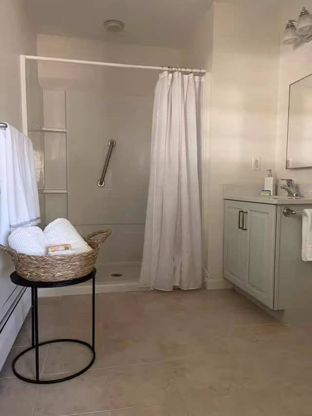 A clean bathroom featuring a walk-in shower with a white curtain and a grab bar inside. To the right is a vanity with a sink, soap dispenser, and a mirror above. On the left side, there is a small round black table holding a woven basket filled with rolled white towels and a bar of soap. A towel hangs on a rack attached to the wall.
