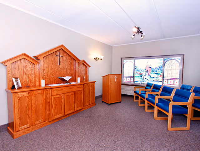 A small chapel room with wooden altar featuring a cross, an open Bible, and candles. To the right, there is a wooden lectern with a cross and a row of blue cushioned chairs. A large stained glass window depicting a religious scene is on the far wall.
