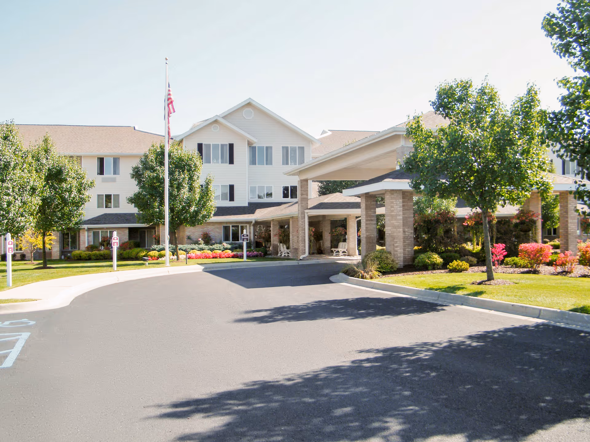 Exterior view of Holiday Genesee Gardens senior living facility showing a multi-story building with white siding and black shutters, a covered entrance with brick pillars, a flagpole with an American flag, well-maintained landscaping with trees and colorful flowers, and a paved driveway.