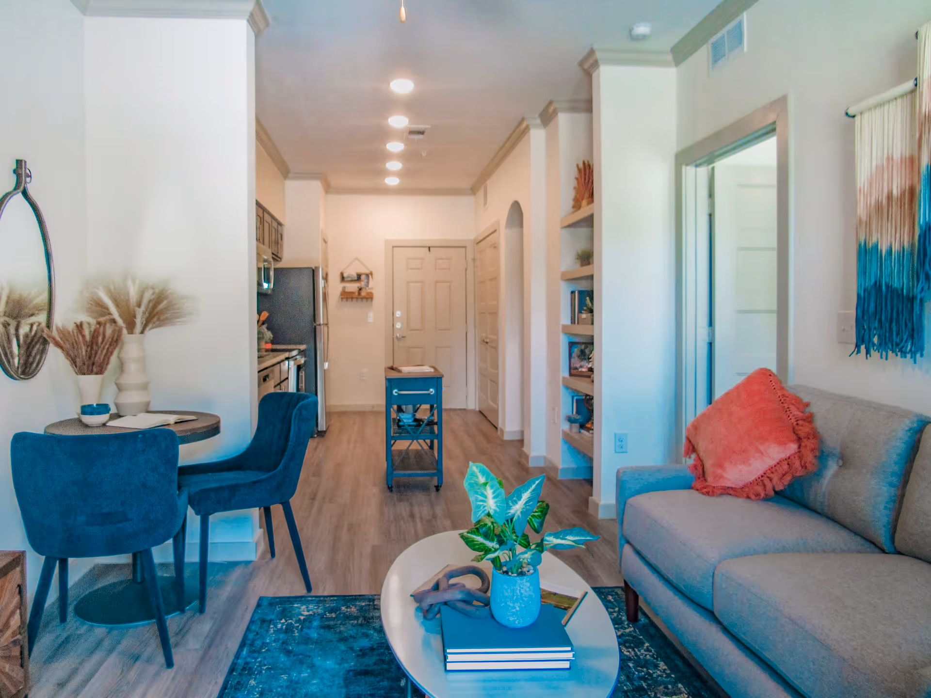 Interior view of a senior living facility apartment showing a small living room with a gray couch and coral pillow, a coffee table with a plant and books, a blue rug, a small dining area with two blue chairs and a round table, and a kitchen area with stainless steel appliances in the background.