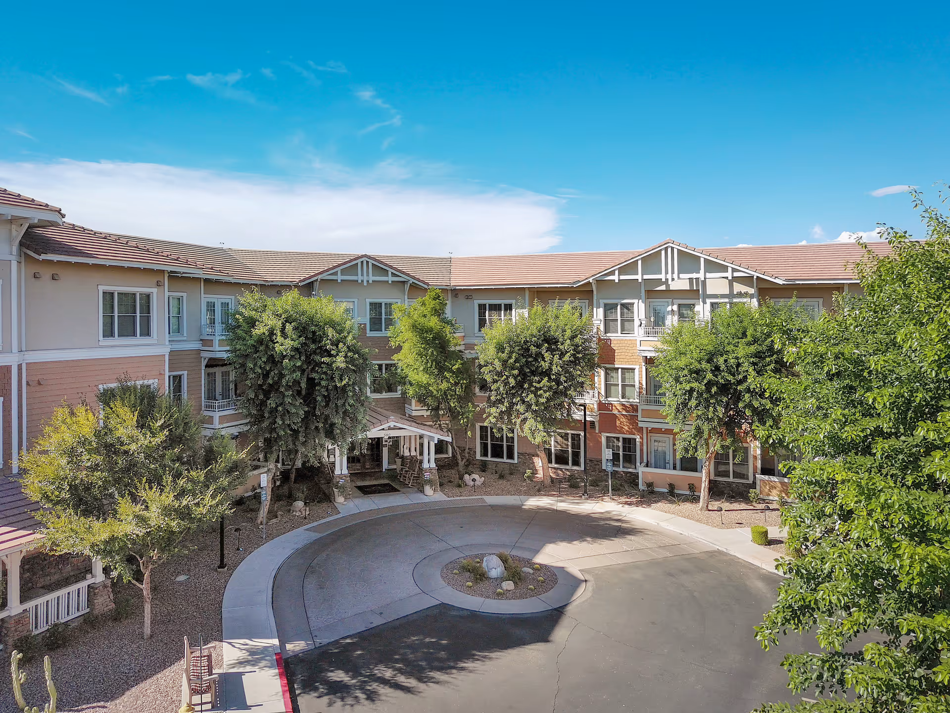 Exterior view of a three-story senior living facility building with a circular driveway and landscaped trees under a clear blue sky.