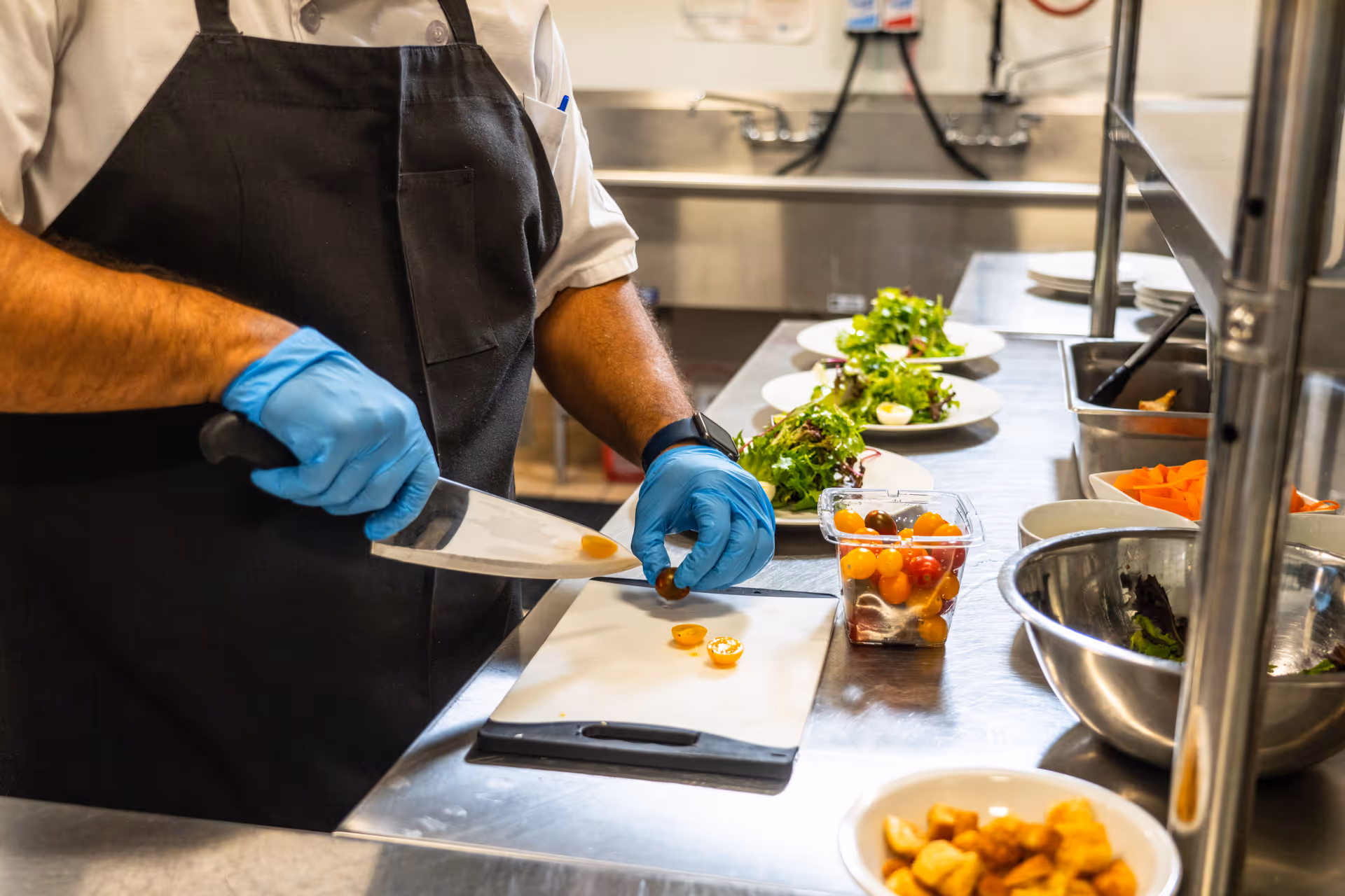 A person wearing a black apron and blue gloves is slicing cherry tomatoes on a cutting board in a kitchen. Several plates with salad and other ingredients are lined up on a stainless steel counter.