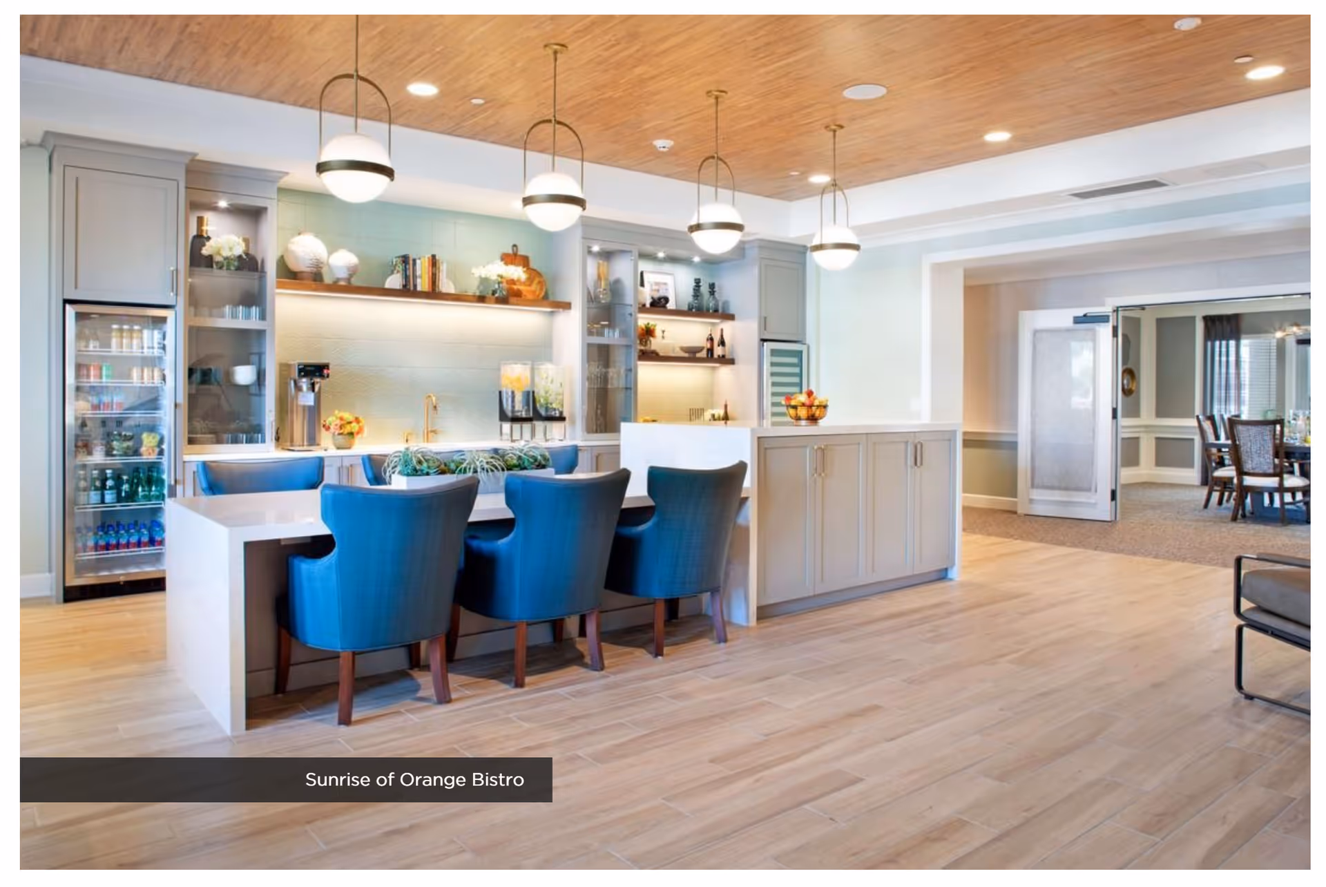 A modern bistro area with a white countertop island surrounded by blue upholstered chairs. The back wall features shelves with decorative items, a beverage dispenser, and a refrigerator stocked with drinks. The ceiling has wooden paneling with hanging globe pendant lights. Adjacent to the bistro is a dining room with a table and chairs visible through an open doorway.