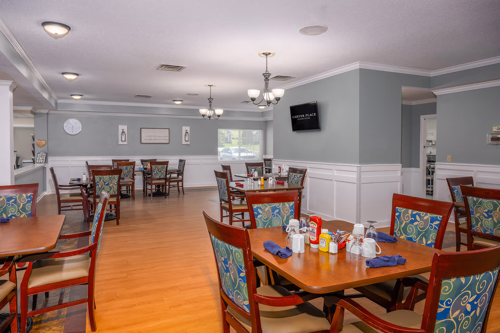 Bright dining room with several wooden tables and patterned chairs set for meals, chandeliers overhead, and a wall-mounted TV reading 'Carter Place'.