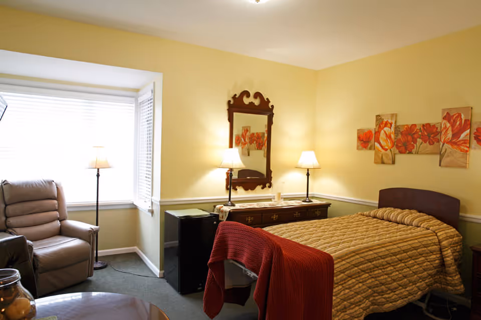 A cozy bedroom with a single bed covered in a beige quilt and a red blanket at the foot. There is a wooden dresser with two lamps and a decorative mirror above it. A comfortable recliner chair is placed near a large window with white blinds. The walls are painted yellow and decorated with floral artwork.