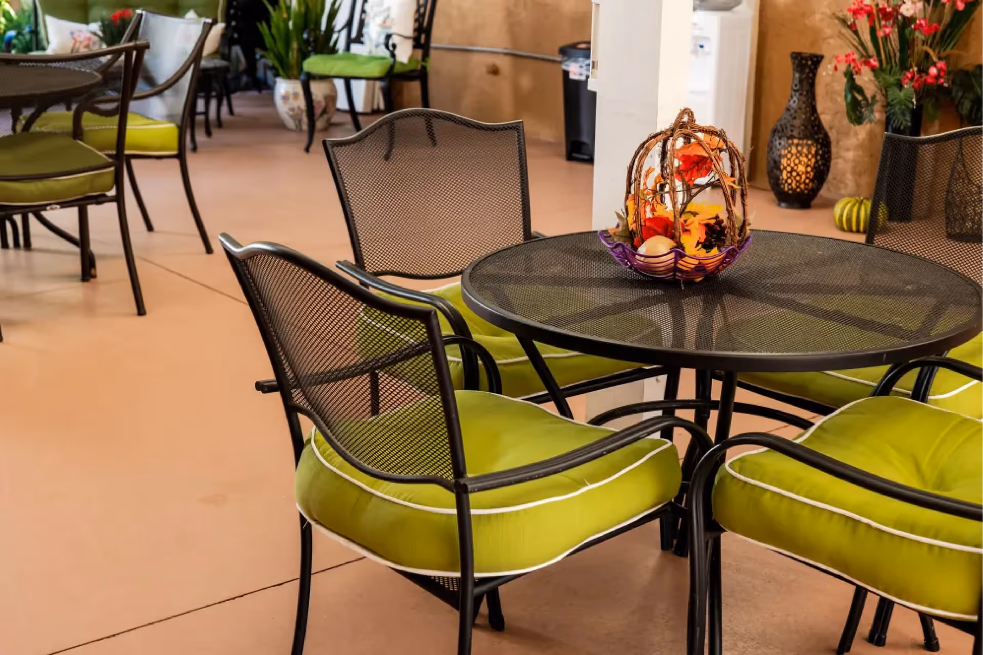 Indoor seating area with black metal mesh table and chairs with green cushions. The table has a decorative centerpiece with autumn-themed items. In the background, there are more chairs and plants in pots against a beige wall.