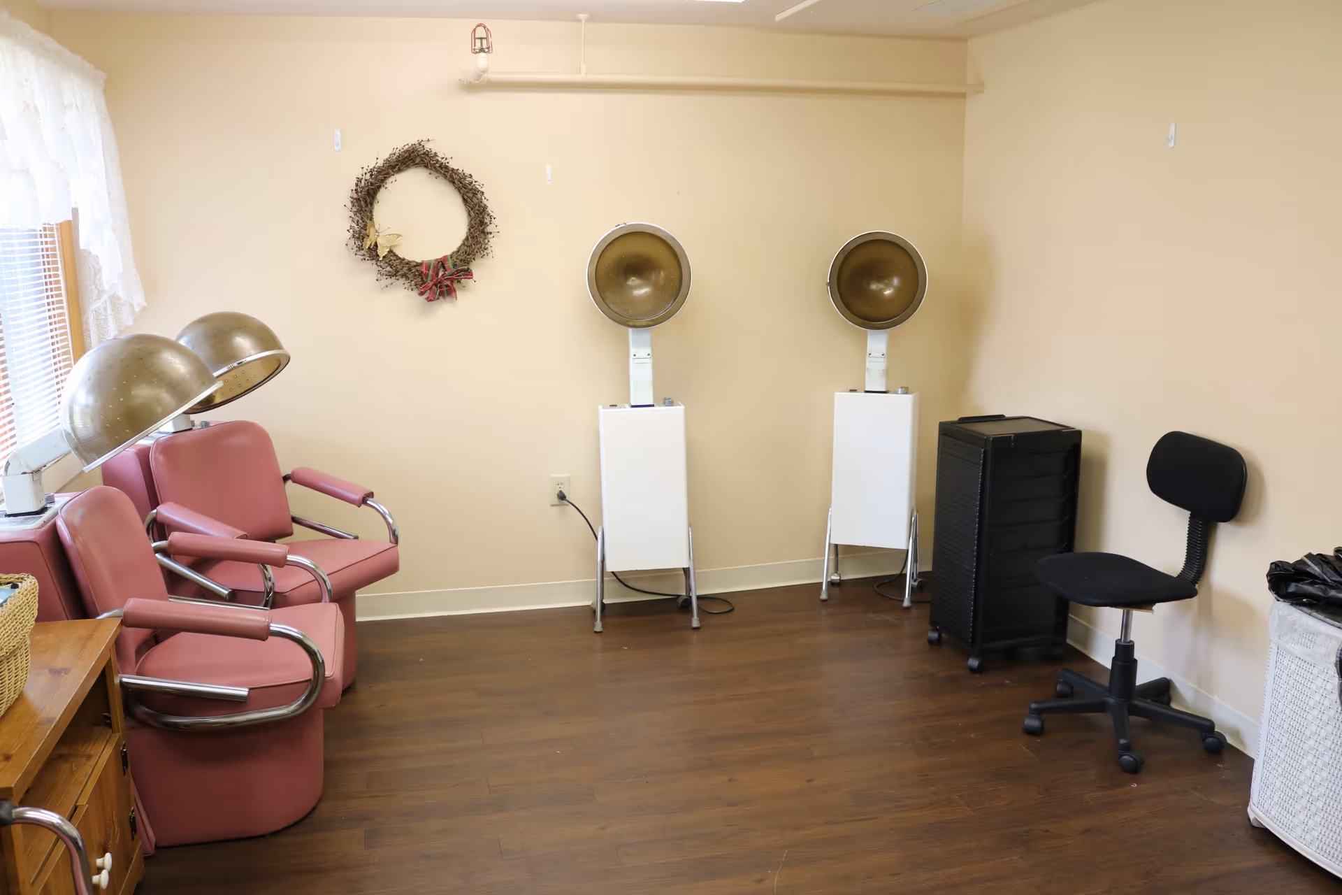 A room with three vintage pink salon chairs equipped with hair dryers, a black rolling chair, a black storage cart, and a wicker basket on a wooden cabinet. The walls are beige, and there is a decorative wreath hanging on the wall. The floor is wooden.