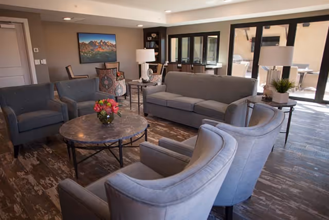 Communal living room with gray sofas and armchairs arranged around a round marble coffee table and large glass doors in the background.
