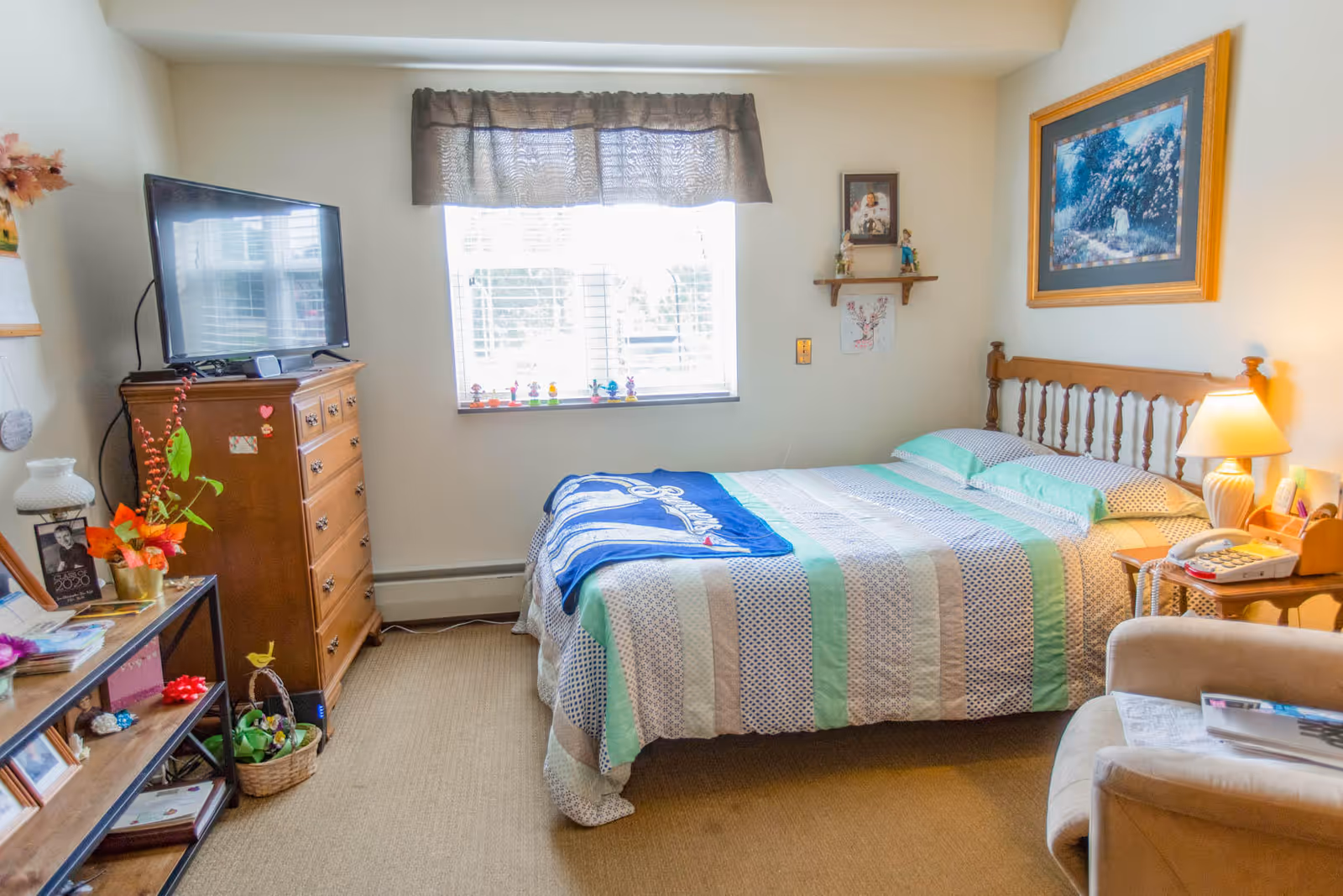 A bright, homey bedroom with a made bed, dresser topped by a TV, a window, nightstand with lamp, and an armchair.