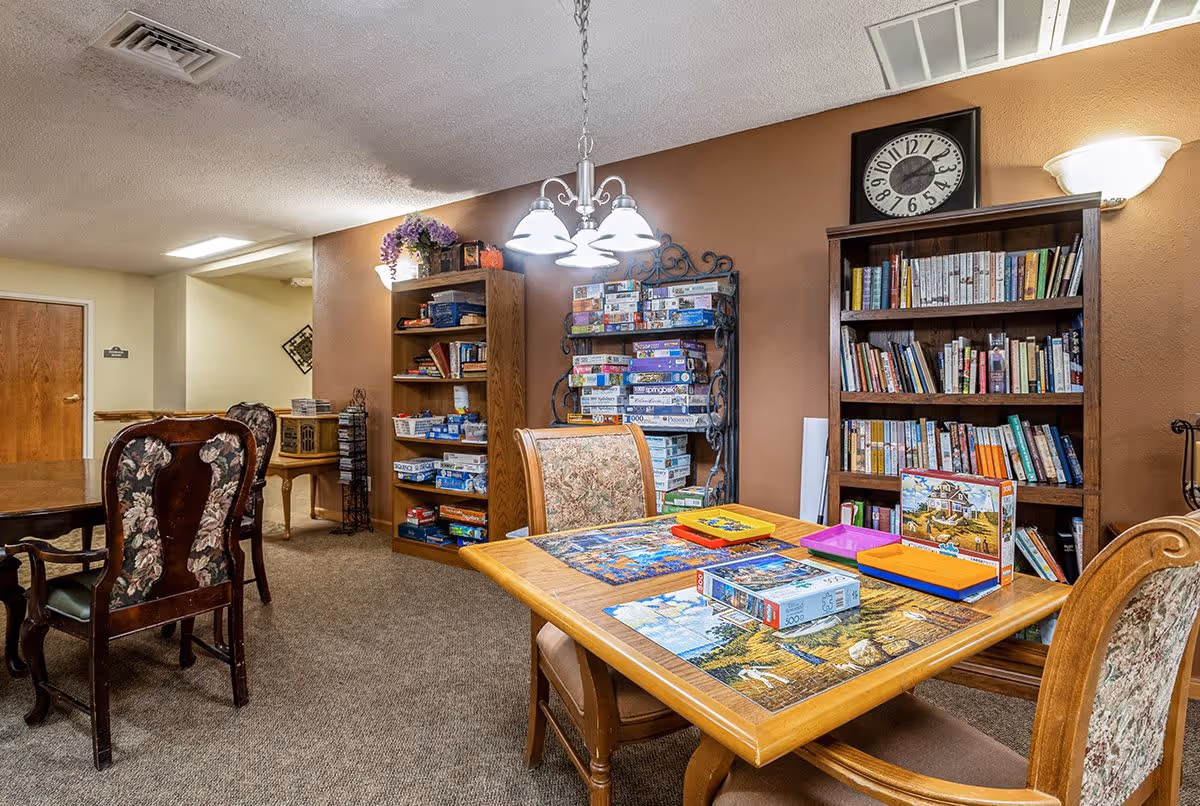 A cozy activity room with wooden tables and chairs, shelves filled with board games and books, a wall clock, and a table set up with puzzle pieces and boxes. The walls are painted brown and beige, and the room is well-lit with ceiling lights and a wall sconce.
