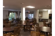 Spacious communal dining room with multiple round tables and wicker chairs, columns, and windows.