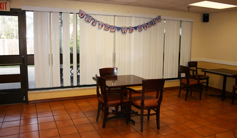 A small dining area with two wooden tables and several chairs on a tiled floor. Vertical blinds cover large windows, and a banner reading 'UNITED WE STAND' is hung across the window. A door with an exit sign is visible on the left side.