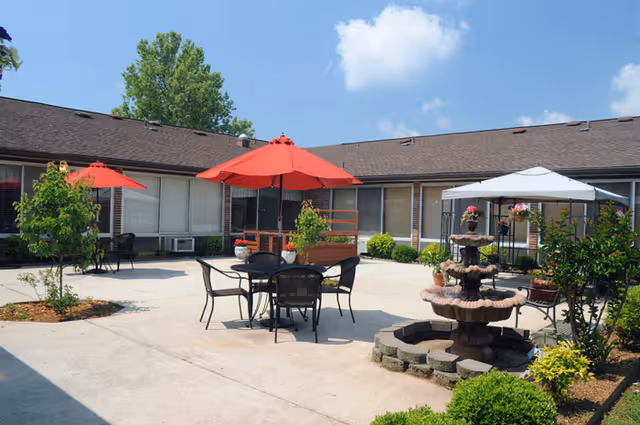 Sunlit courtyard at a nursing facility with patio tables and red umbrellas, a decorative fountain, and surrounding single-story building.
