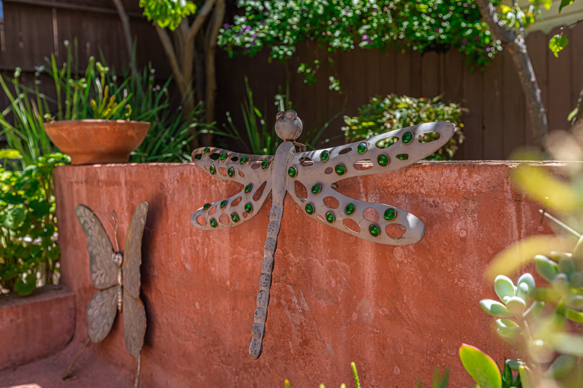 A decorative metal dragonfly with green glass accents mounted on a reddish stone wall in a garden area, with a metal butterfly decoration and various green plants in the background.