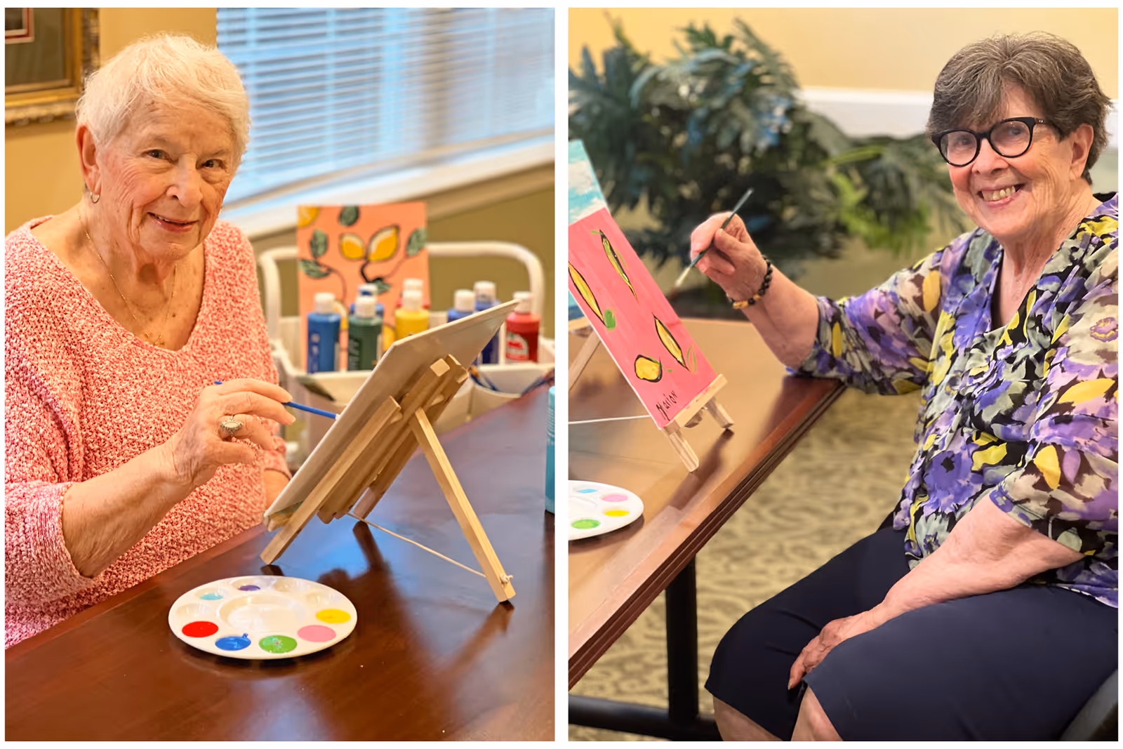 Two elderly women sitting at a table painting on canvases placed on small easels. One woman wears a pink sweater and the other wears a purple floral blouse. Various paint bottles and palettes with different colors are visible on the table.