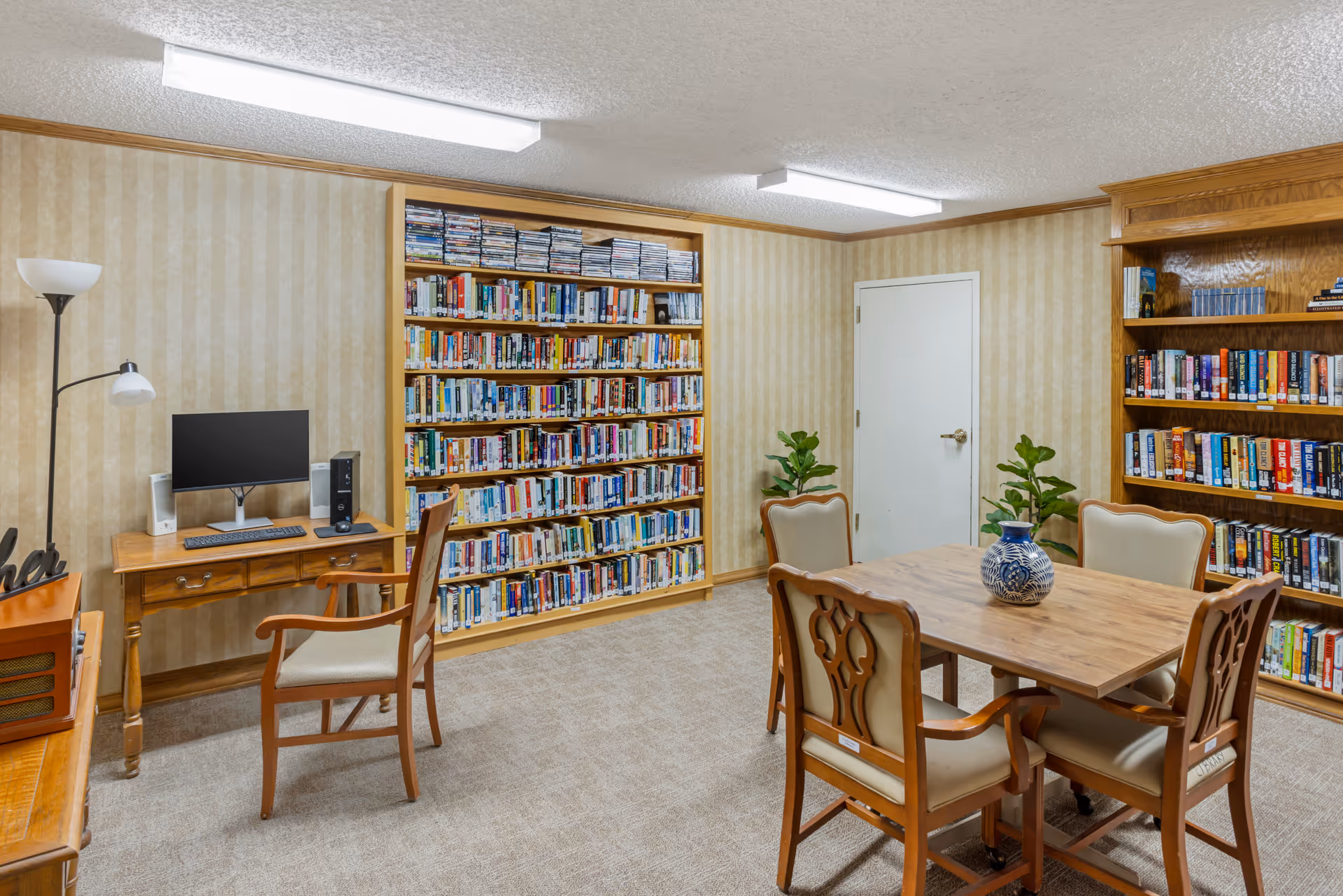 A cozy interior library/activity room with bookshelves lining the walls, a table and chairs, and a computer workstation.