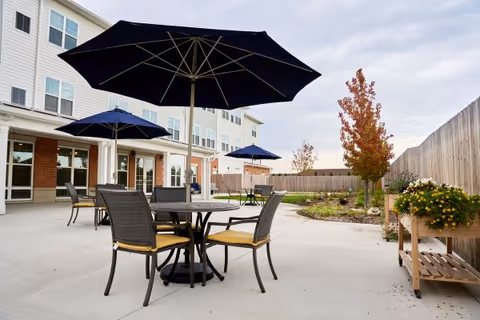 Outdoor patio area at a senior living facility with round tables, chairs with yellow cushions, and large navy blue umbrellas. The patio is adjacent to a multi-story building with large windows. There are small trees and plants along the fenced perimeter of the patio.