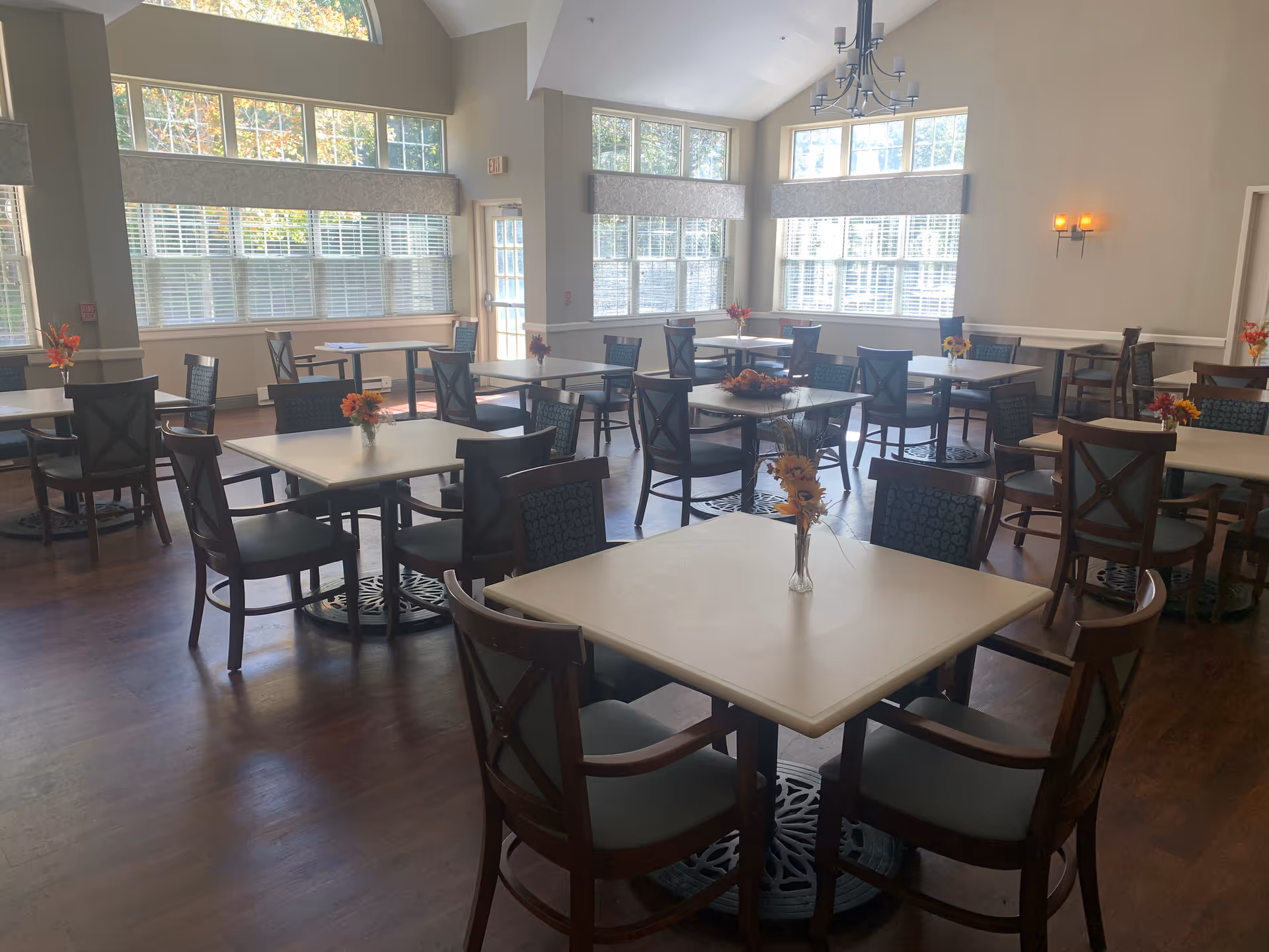 Bright dining room with multiple square tables and chairs, small floral centerpieces, large windows, and hardwood floors.