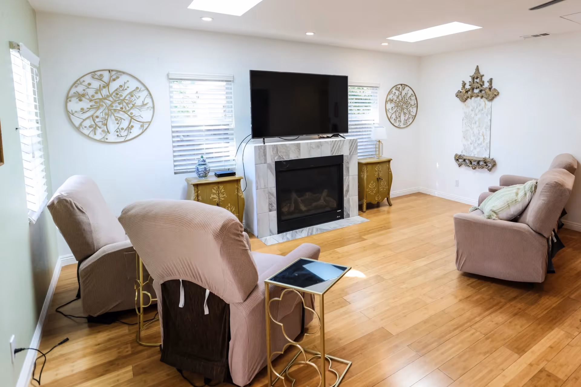 A bright living room with wooden flooring, two windows with white blinds, a wall-mounted flat-screen TV above a tiled fireplace, two gold side tables with decorative items, and four beige armchairs arranged around the fireplace. The walls are white with decorative wall art and a mirror.