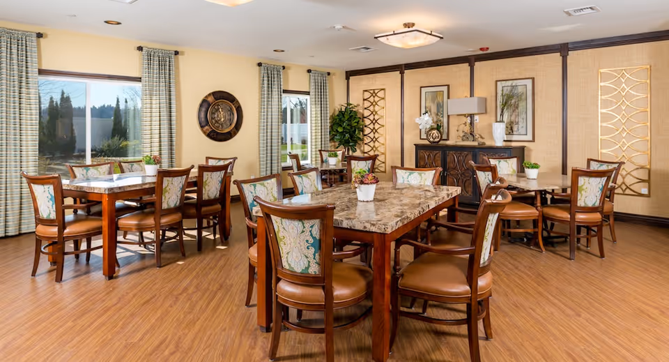 A bright dining room with multiple wooden tables and chairs featuring patterned upholstery. The room has large windows with striped curtains, wooden flooring, decorative wall art, and a sideboard with a lamp and plants.