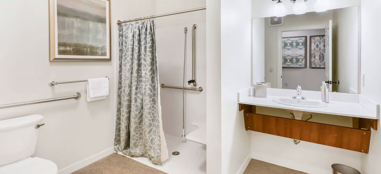 A clean and accessible bathroom featuring a white toilet, a shower area with a patterned curtain and grab bars, a large mirror above a white sink with a wooden panel underneath, and a towel hanging on a grab bar. The walls are light-colored and there is a framed artwork above the toilet.