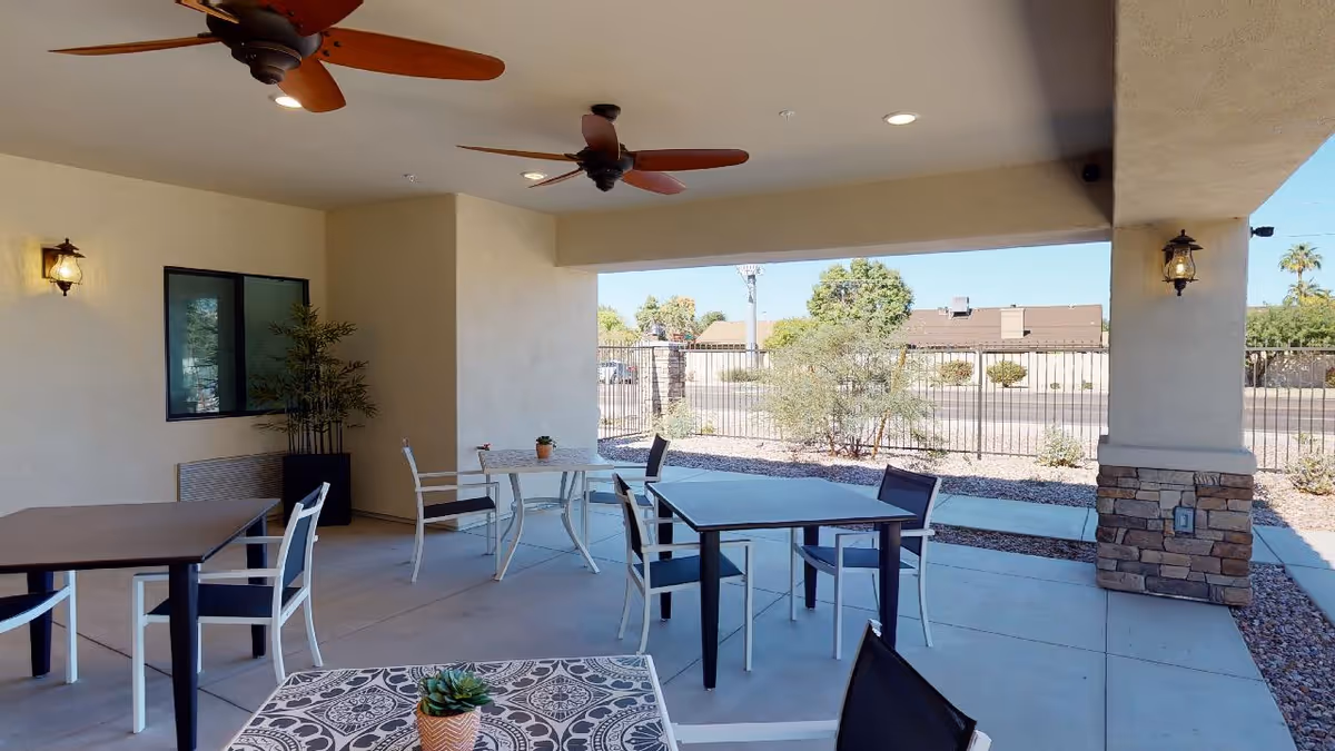 Covered outdoor patio area with several tables and chairs arranged for seating. Ceiling fans are mounted on the ceiling, and there are wall-mounted lantern-style lights. The patio overlooks a fenced area with some shrubs and trees, and a sidewalk runs along the edge.