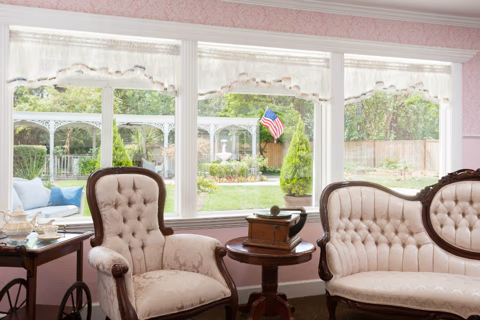 A cozy sitting area with vintage upholstered armchair and loveseat in front of large windows overlooking a garden with an American flag, white gazebo, and a fountain.