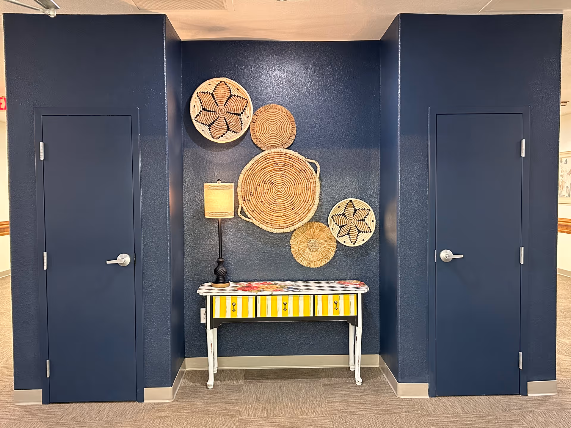 Hallway alcove with navy walls featuring woven basket wall decor, a small striped accent table with a lamp between two closed navy doors.