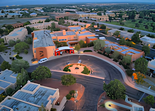 Aerial view of The Montecito of Santa Fe senior living facility showing multiple adobe-style buildings arranged around a circular driveway with surrounding greenery and trees under a clear sky.