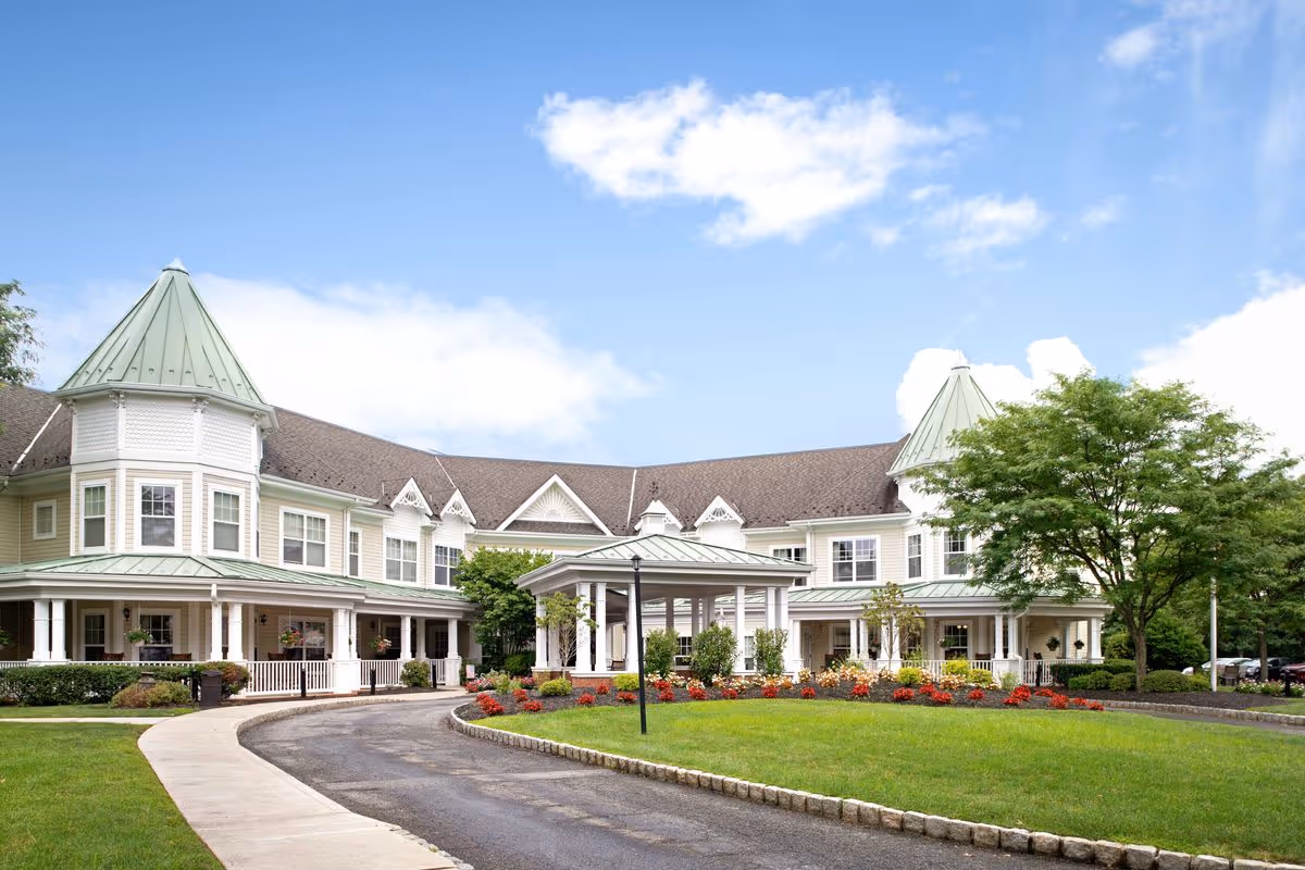 Exterior view of a large, two-story senior living facility with light-colored siding and green metal roofs on turret-like structures. The building is surrounded by well-maintained landscaping including green grass, trees, and colorful flower beds. A curved driveway leads to a covered entrance in front of the building under a partly cloudy blue sky.
