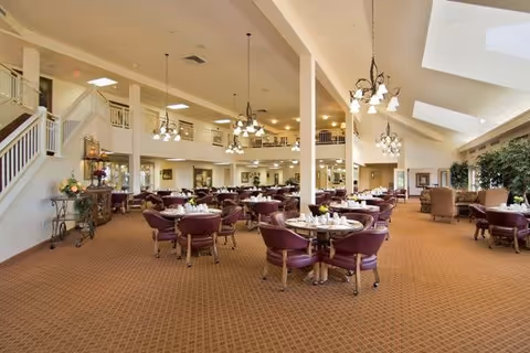 Spacious dining room with multiple round tables set with white tableware and purple cushioned chairs. The room features high ceilings with hanging chandeliers, large windows allowing natural light, and a staircase leading to an upper level. There are also some lounge chairs and plants along the walls.
