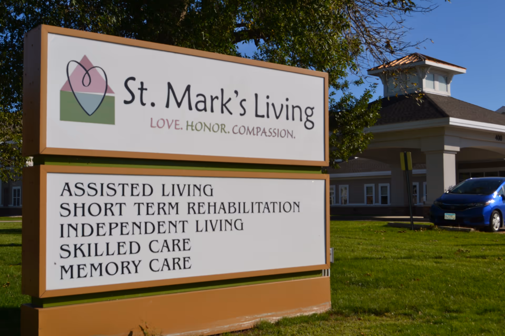 Outdoor sign for St. Mark's Living assisted living facility with services listed including assisted living, short term rehabilitation, independent living, skilled care, and memory care. The building and a blue car are visible in the background under a clear blue sky.