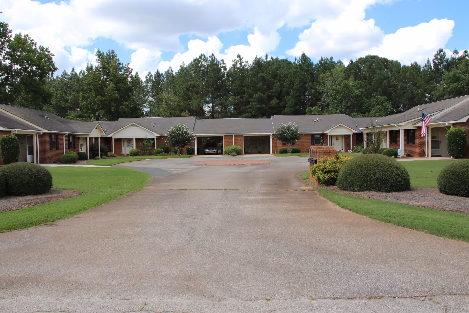 Exterior view of a single-story brick senior living facility with a circular driveway, neatly trimmed bushes, green lawns, and trees in the background under a partly cloudy sky.