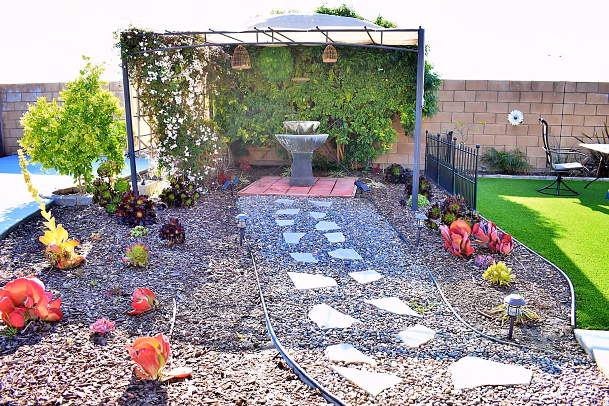 A landscaped outdoor garden area with a stone pathway leading to a two-tiered water fountain under a metal pergola covered with green vines and white flowers. The garden is bordered by mulch with various small plants and succulents, and there is a green artificial turf area with outdoor chairs and a table to the right, enclosed by a low black metal fence.