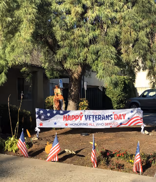 Outdoor scene at Medford Rehabilitation & Nursing Center featuring a large tree with a carved wooden soldier statue beneath it. In front of the statue is a banner that reads 'Happy Veterans Day Honoring All Who Served' decorated with stars and an American flag design. Small American flags are planted in the ground along the sidewalk near the banner.