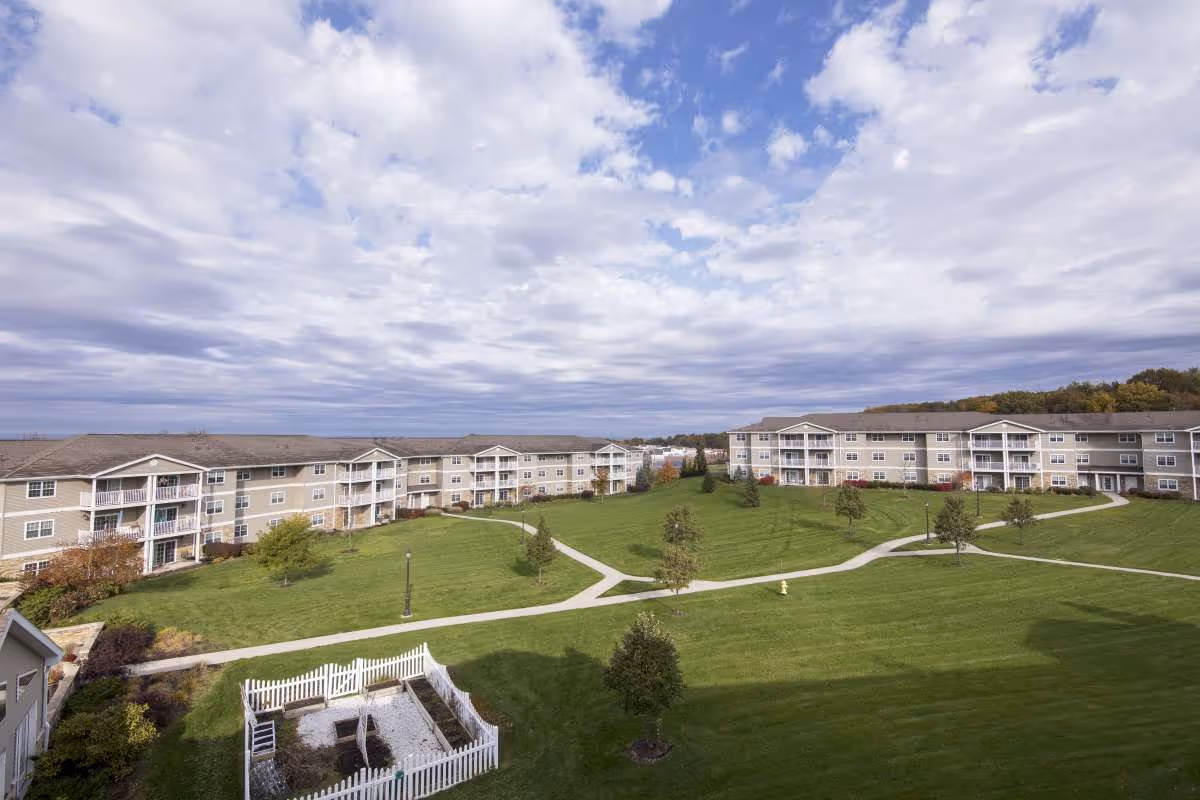 View of a senior living facility with multiple three-story beige buildings surrounding a large green lawn with paved walking paths, small trees, and a white fenced garden area under a partly cloudy sky.