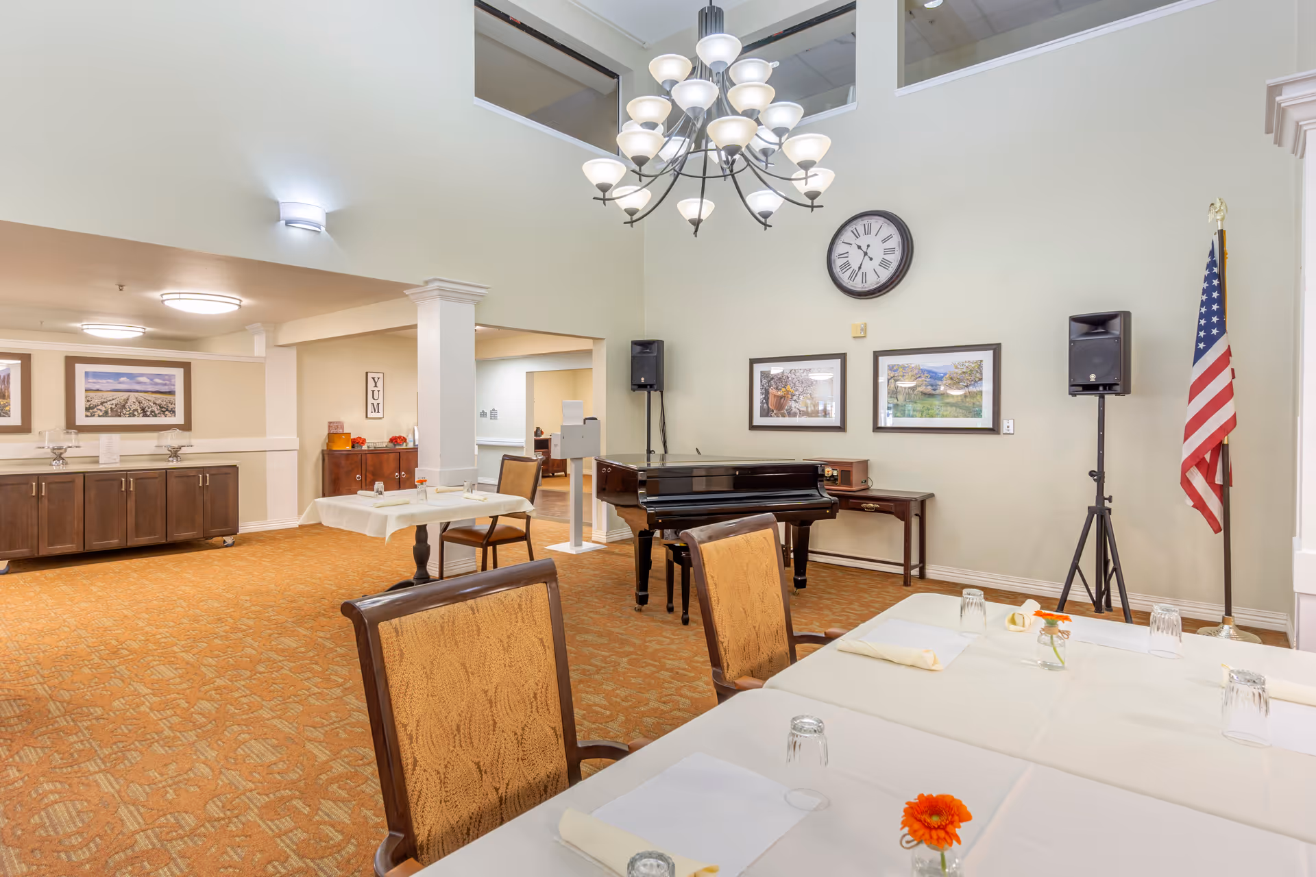 Bright communal dining/activity room with tables set, a grand piano, chandelier, wall clock and an American flag.